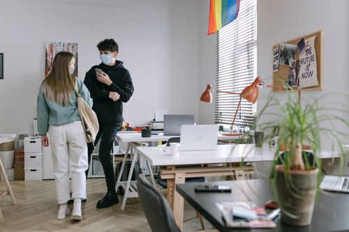 Free Two colleagues in masks interacting in a modern, light-filled office space with open workstations. Stock Photo