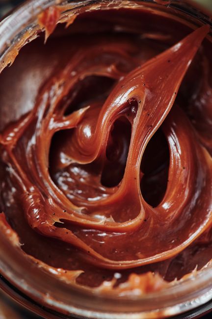 A glass jar of amber cajeta with a spoon lifting a thick ribbon of the caramel, showing its glossy, pourable texture.