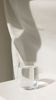 A minimalist composition featuring a glass of water on a white surface with sunlight and shadows.