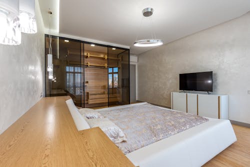 Bed with pillows placed against wall with TV on cupboard in spacious bedroom and glowing chandelier with wardrobe in apartment