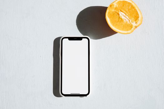 A smartphone with a blank screen next to an orange slice on a light surface, perfect mockup.