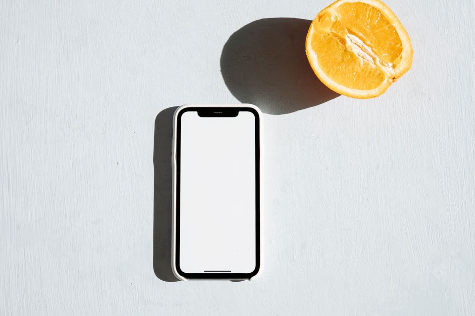 A smartphone with a blank screen next to an orange slice on a light surface, perfect mockup.