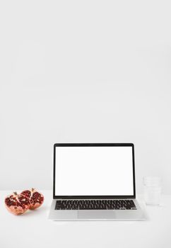 Stylish minimalist laptop mockup with a pomegranate and glass of water on a white desk.