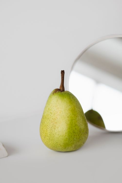 Free A fresh green pear stands in focus on a clean white surface, reflecting in a nearby mirror. Stock Photo