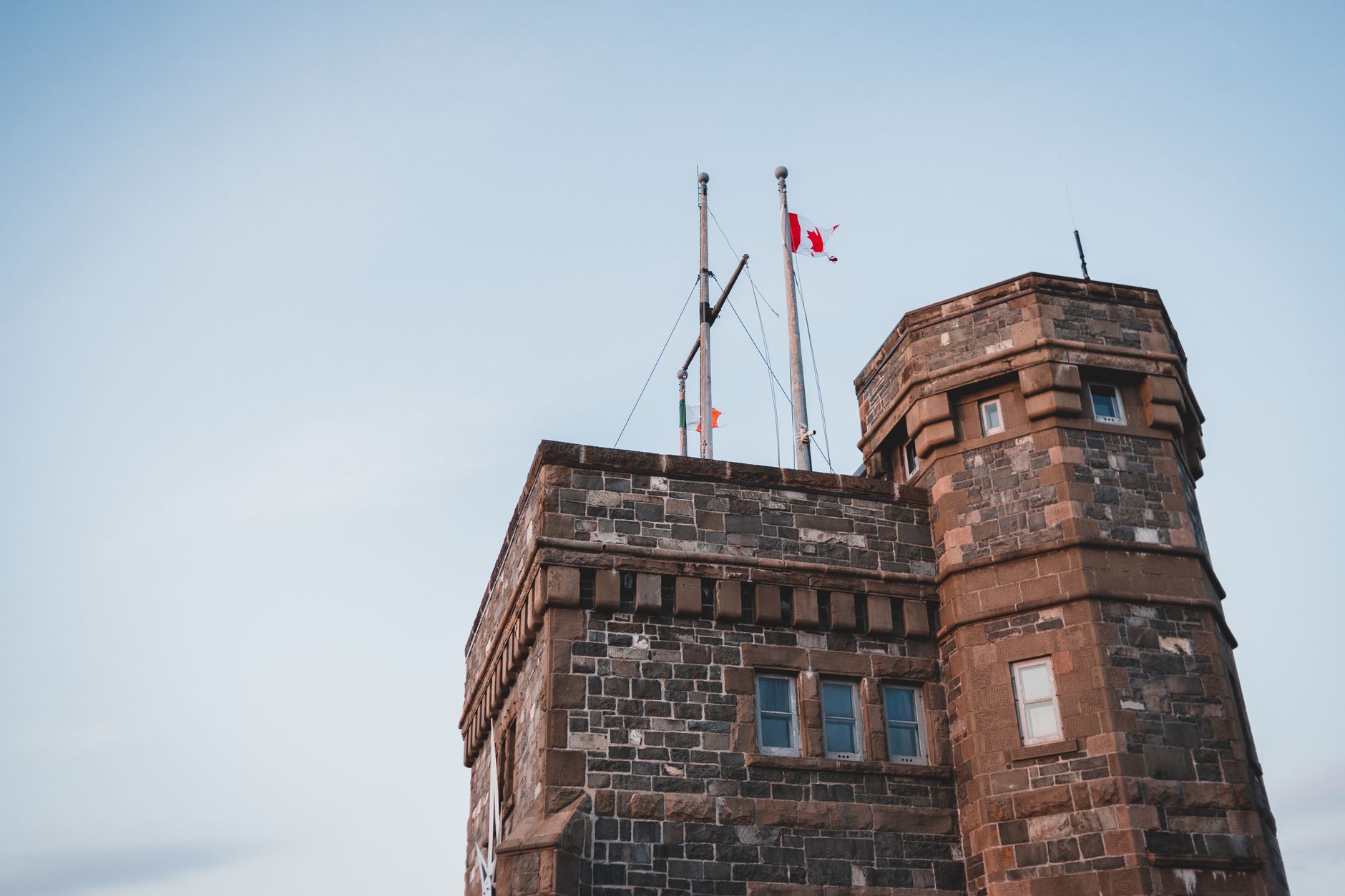 From below of flag of Canada on roof of aged stone Cabot Tower against cloudy blue sky in St Johns