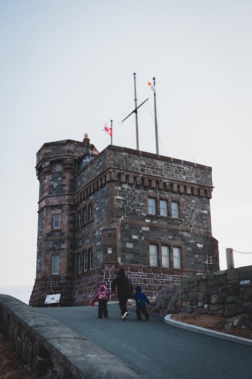 Back view of unrecognizable man holding hands of children and walking on asphalt road towards Cabot Tower located on Signal Hill against cloudy sky