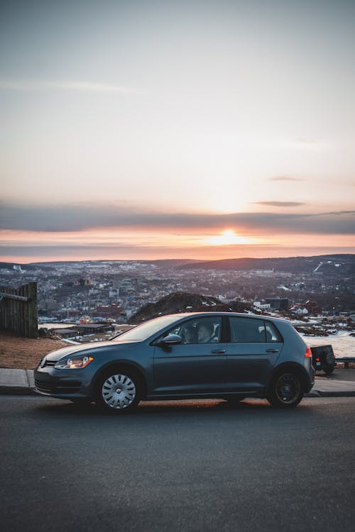 Anonymous travelers in car parked on asphalt road with picturesque city view against sunset sky