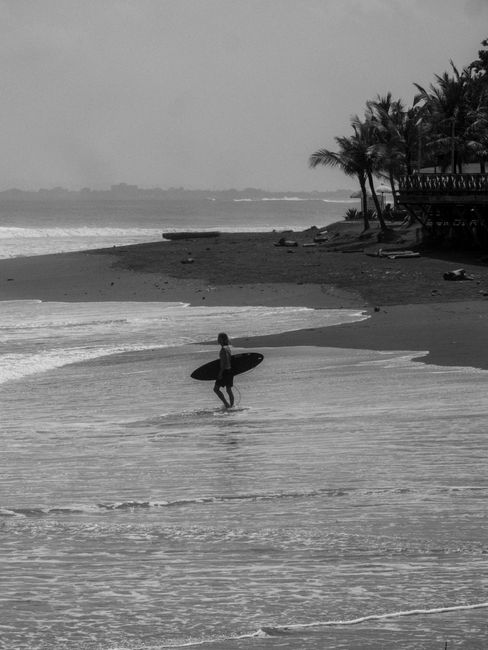Black and white photo of a surfer carrying a surfboard along the beach in Bali, Indonesia.