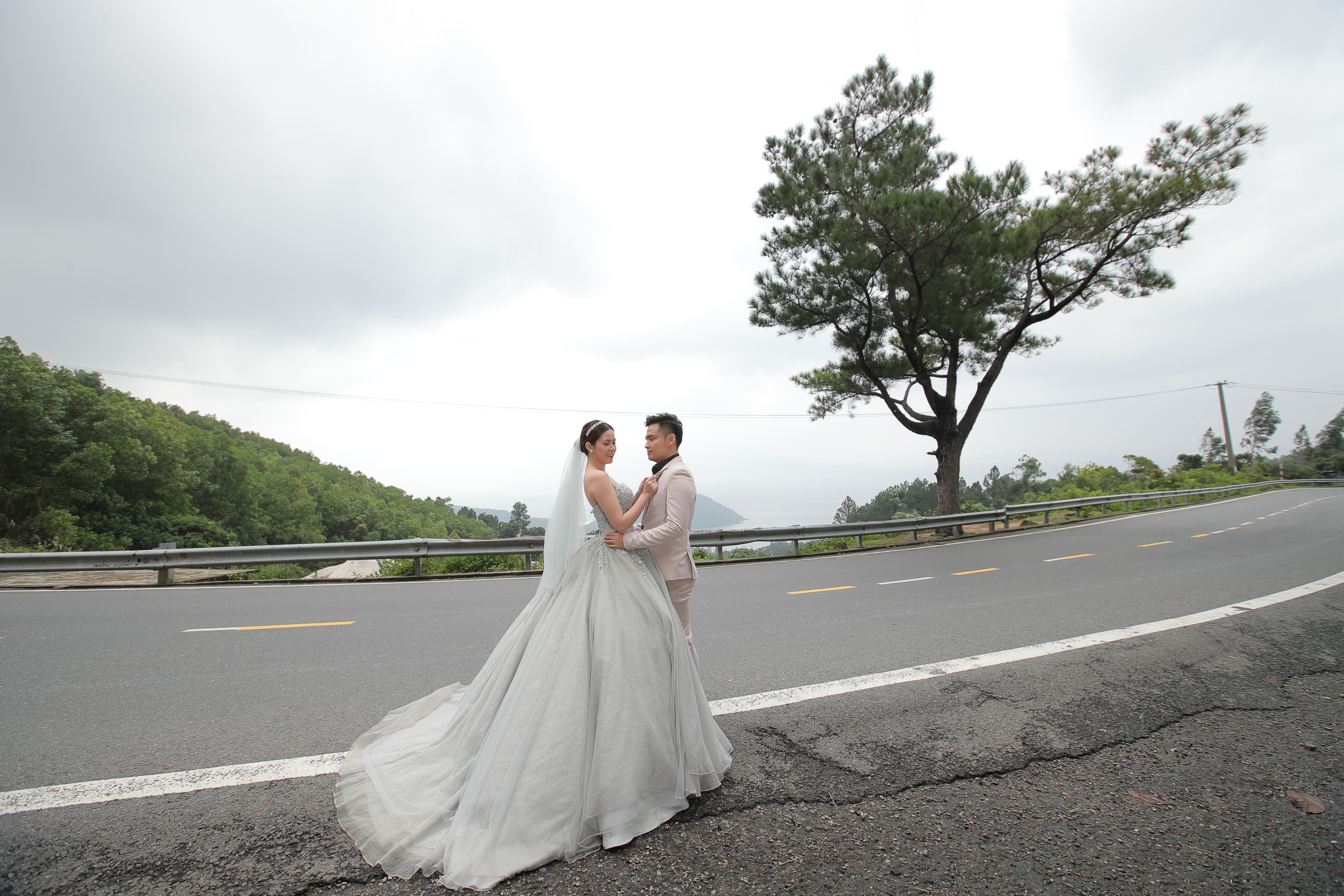 A bride and groom embracing on a scenic road in Thua Thien Hue, Vietnam, under a cloudy sky.