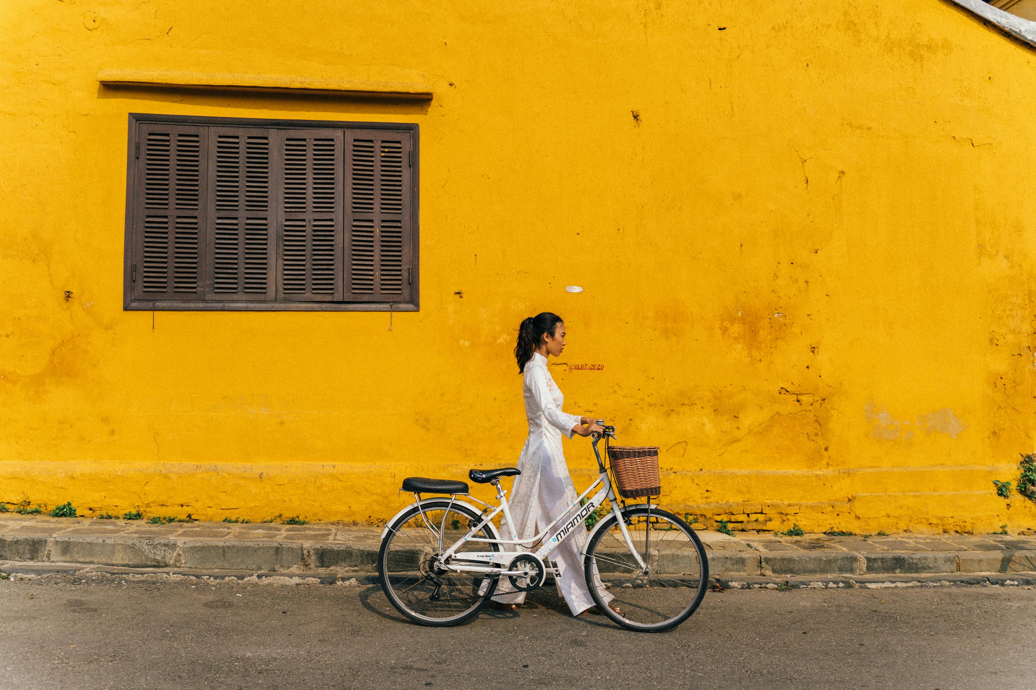 Asian woman in traditional ao dai walking with a bicycle past a bright yellow wall.