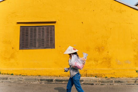A woman in traditional dress walks by a vibrant yellow wall in Hội An, Vietnam, during the day.
