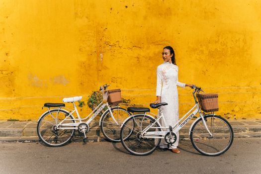 A woman in traditional Ao Dai stands with bicycles against a vibrant yellow wall outdoors.