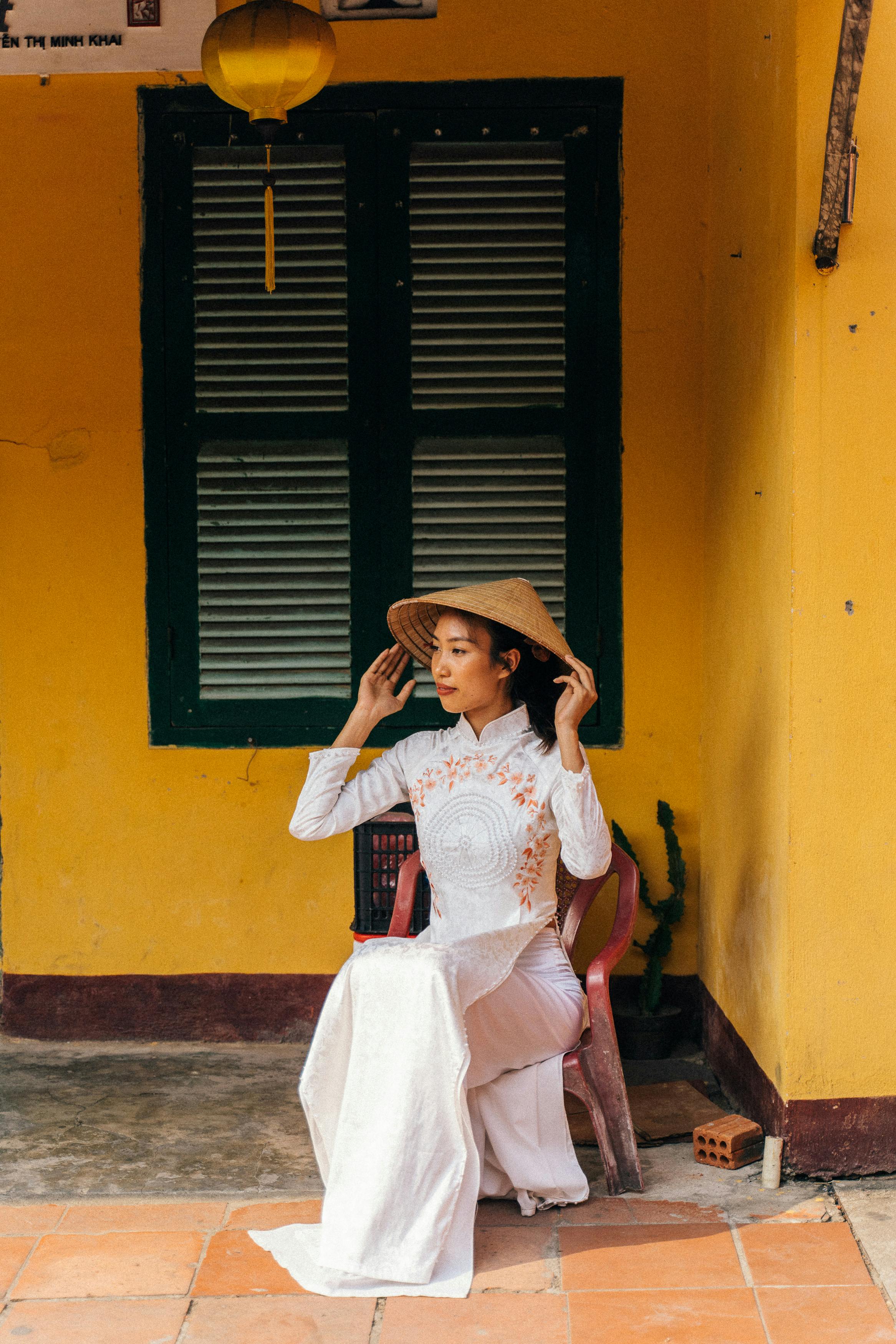 Elegant Vietnamese woman in a traditional ao dai seated outdoors, holding a conical hat.
