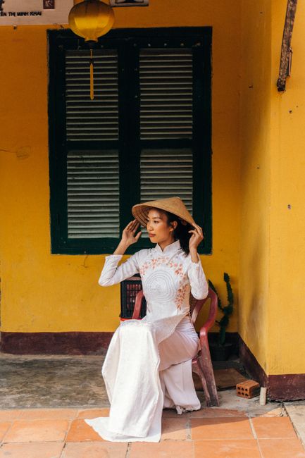 Elegant Vietnamese woman in a traditional ao dai seated outdoors, holding a conical hat.