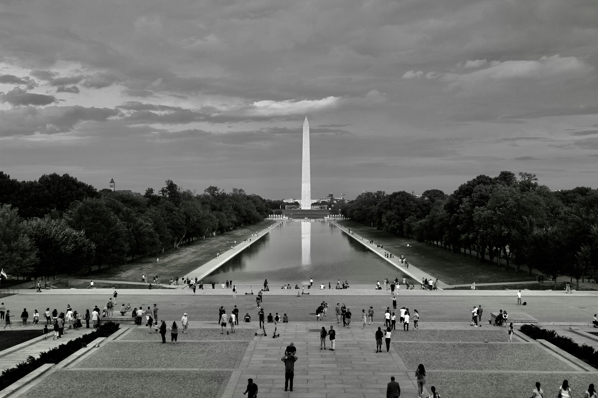 A black and white image of the Washington Monument with people at the Reflecting Pool in Washington, DC.