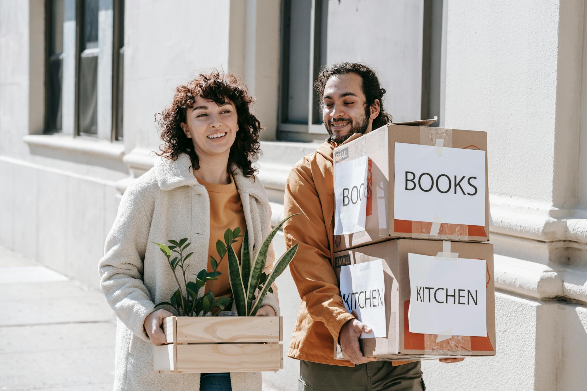 A smiling couple carrying boxes and plants while moving into a new urban home.