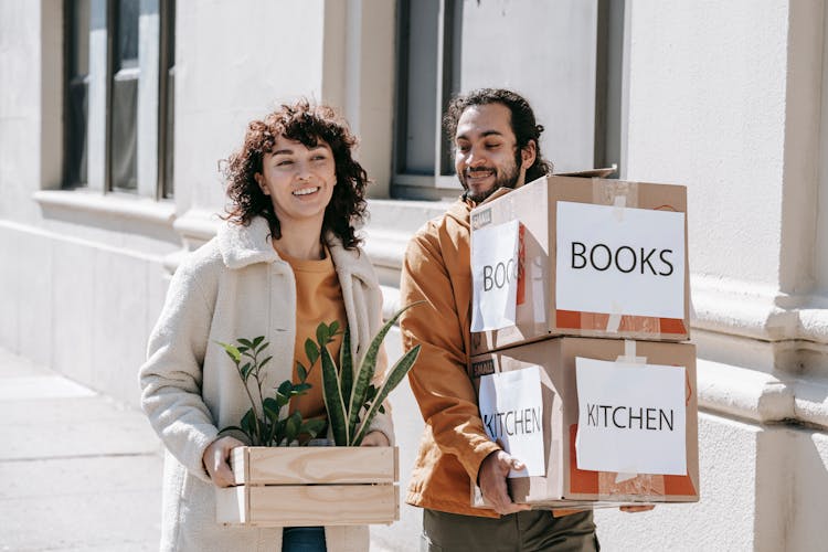 A Couple Moving Out Carrying Boxes And Plants