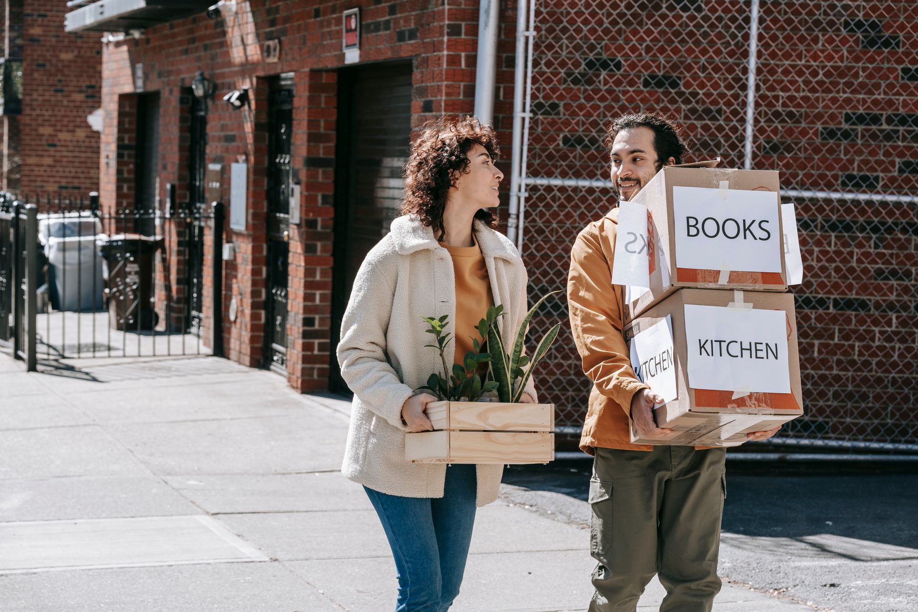 A couple happily moving to a new home carrying boxes and a plant on a sunny day.