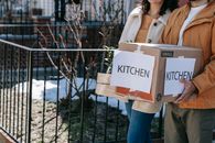 A couple carrying labeled kitchen boxes while moving into a new home outdoors.