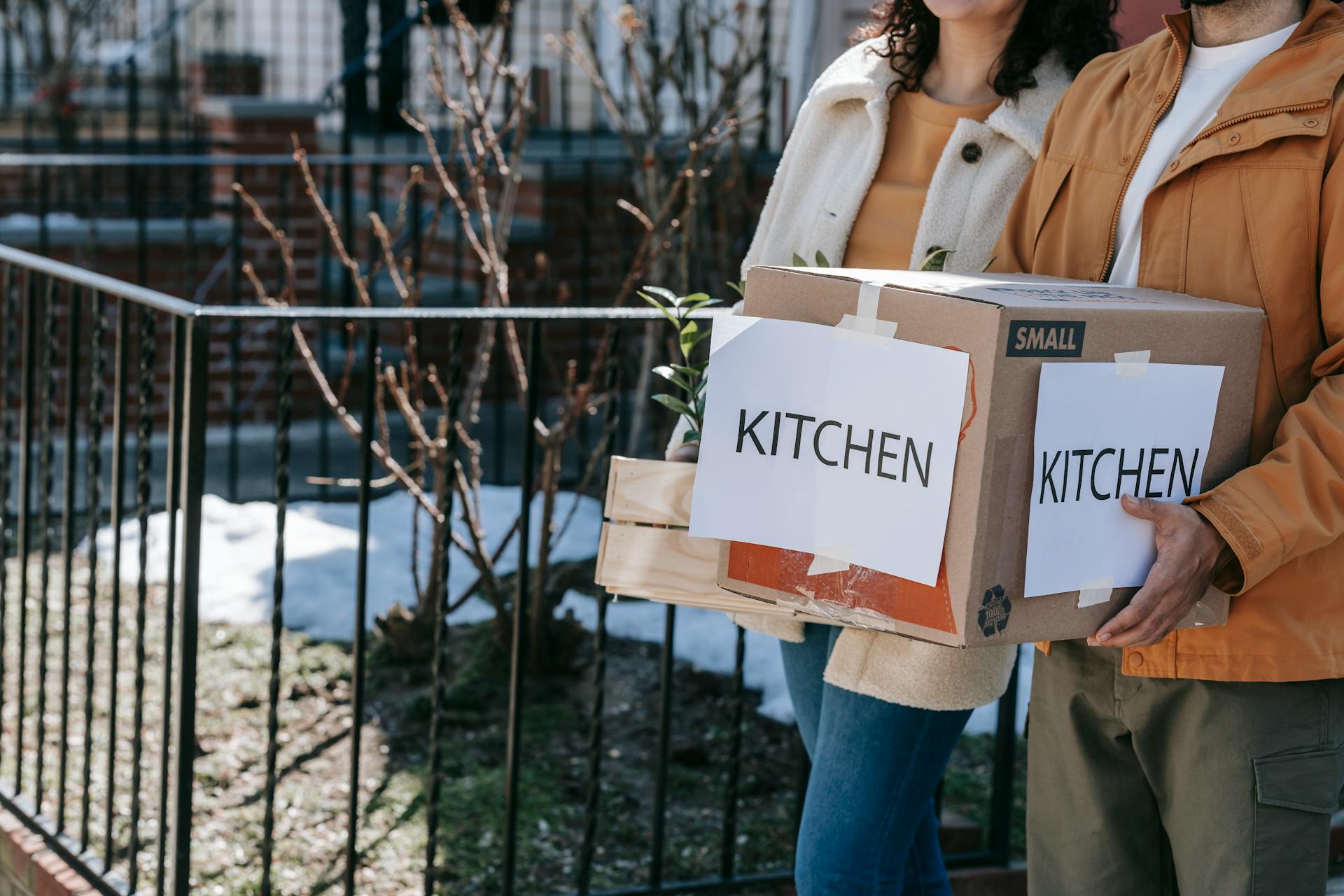 A couple carrying labeled kitchen boxes while moving into a new home outdoors.