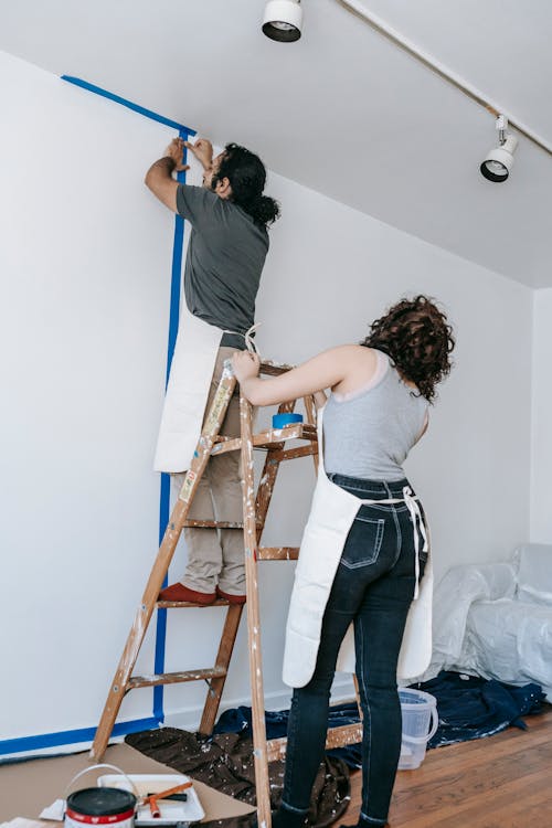 Man Putting Tape On Wall With Woman Holding The Stepladder