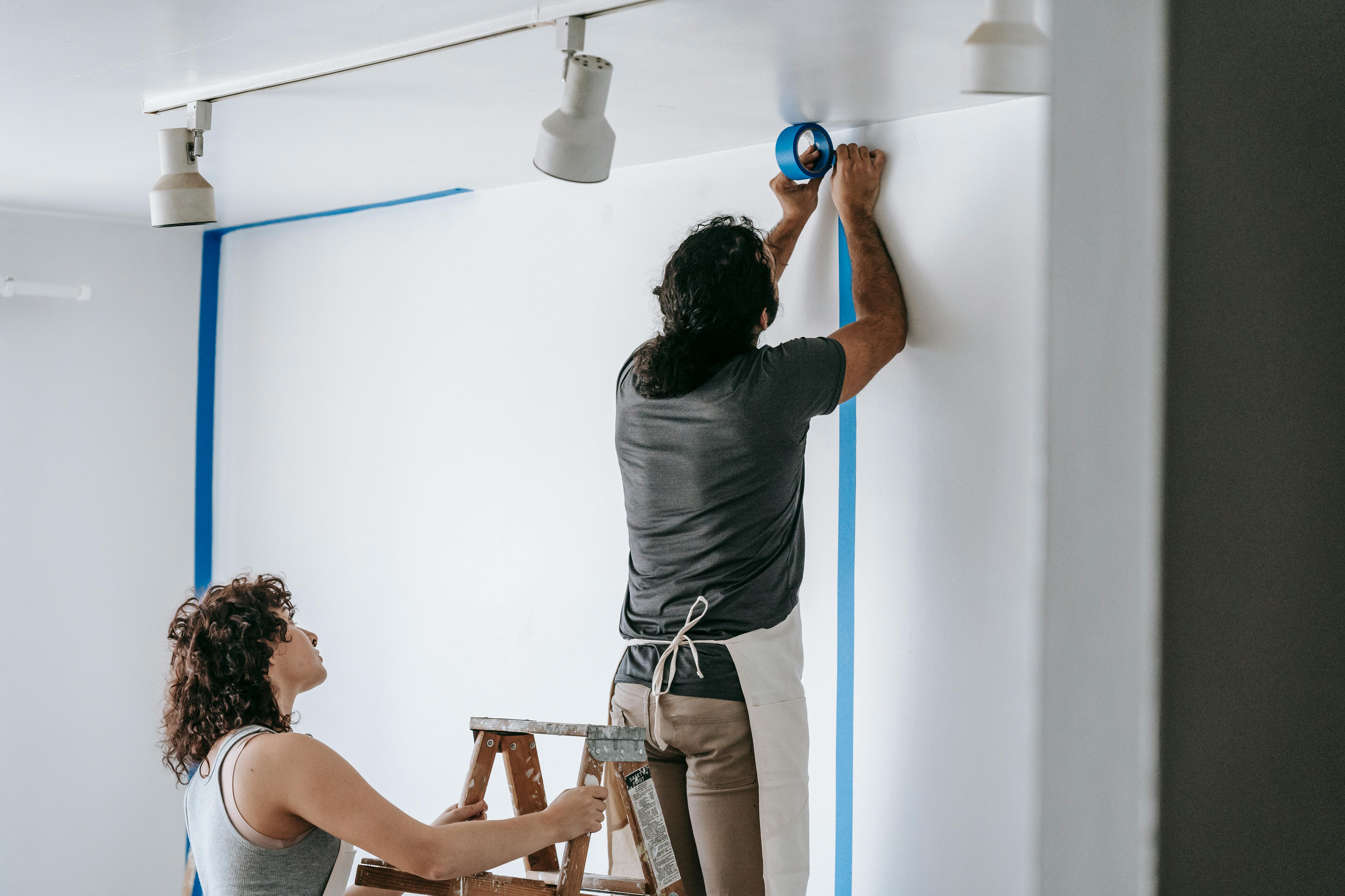 man putting tape on wall with woman holding the stepladder