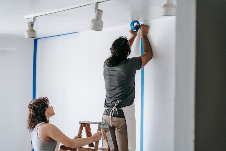 Man Putting Tape On Wall With Woman Holding The Stepladder