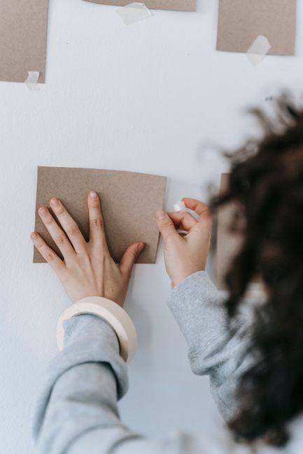 Crop anonymous female with adhesive tape sticking rectangular shaped carton piece on white wall at home
