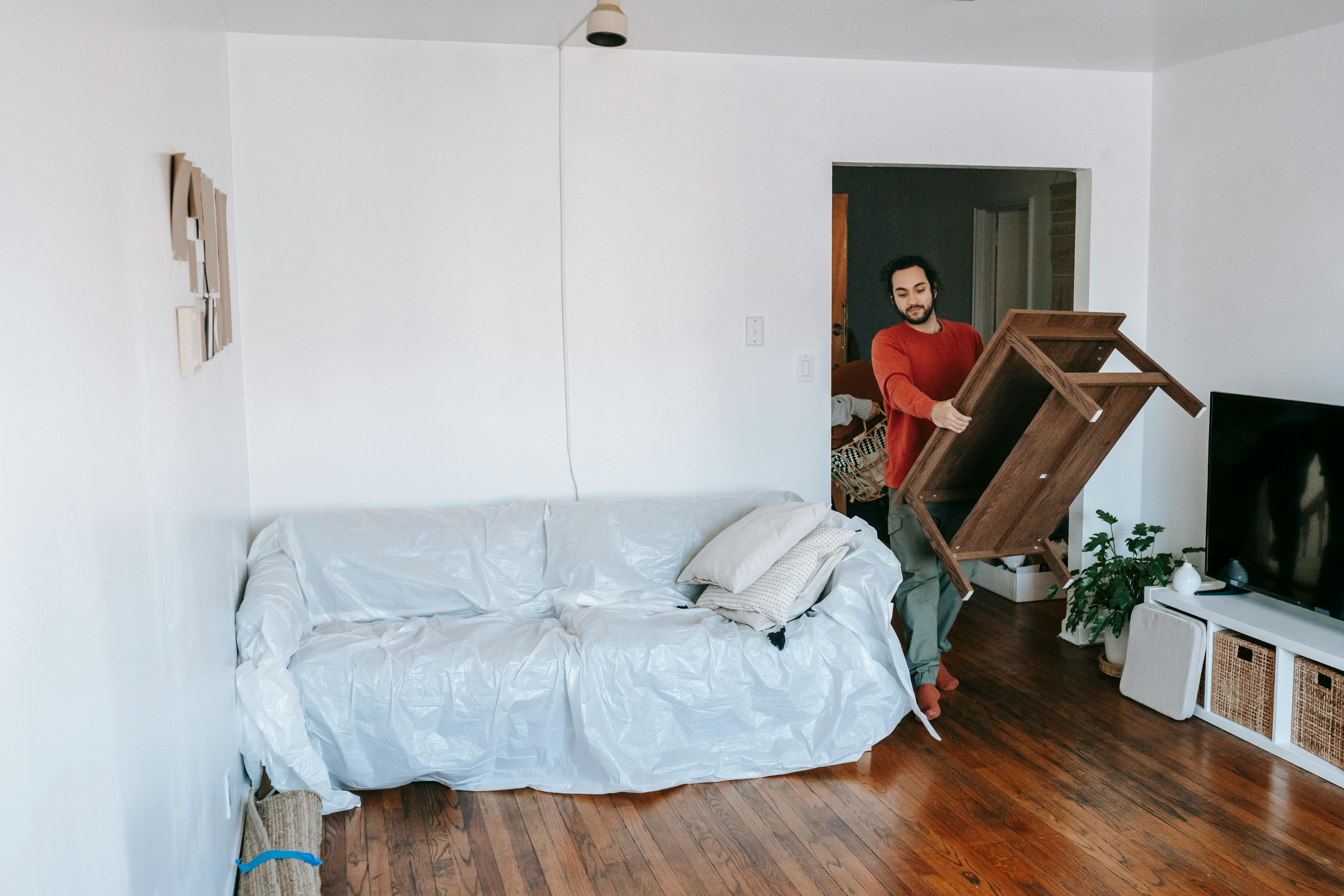 Free Man lifting a wooden table in a bright living room with wrapped sofa, preparing for a move. Stock Photo