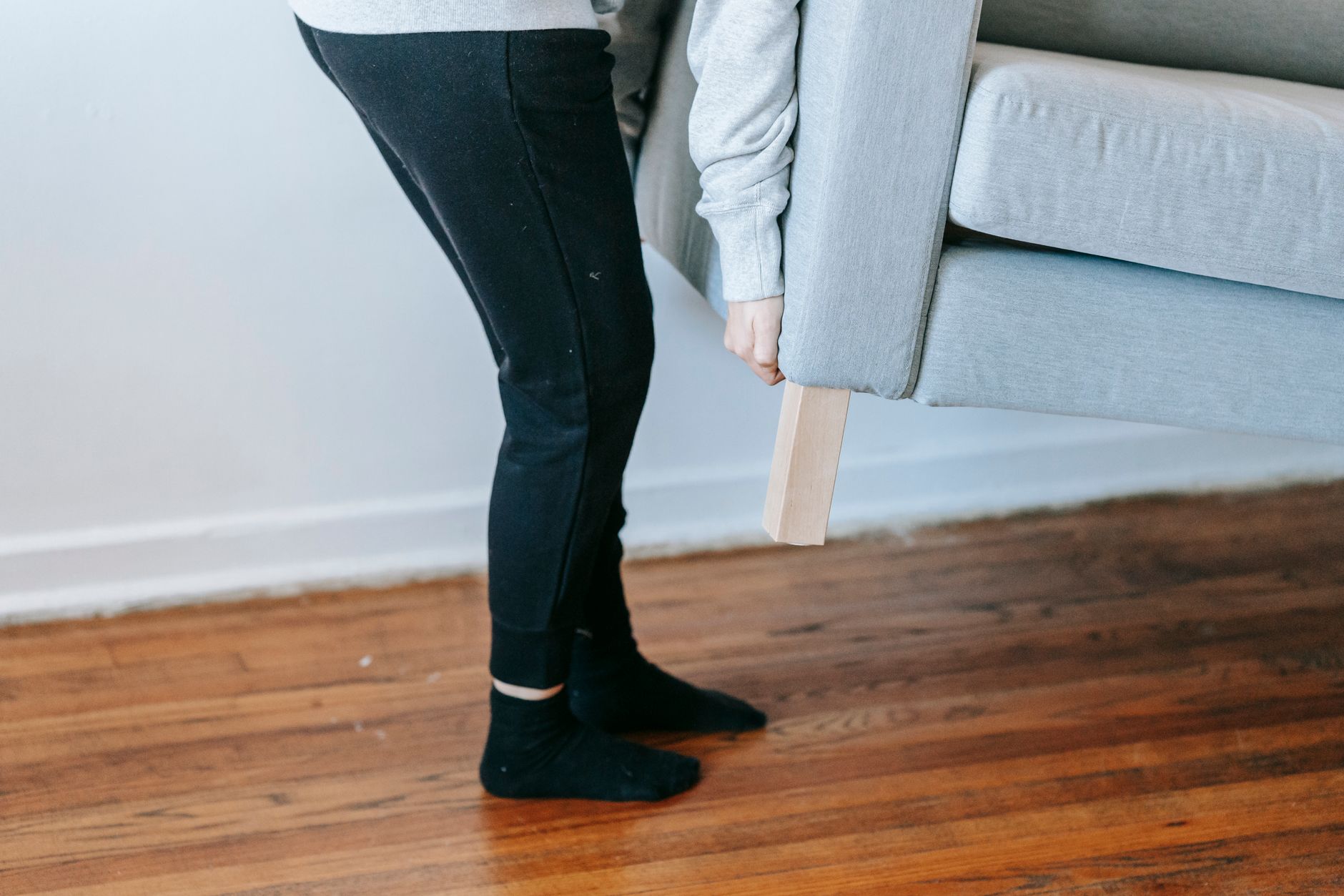 A person lifting a couch in a minimalist living room with wooden floors.