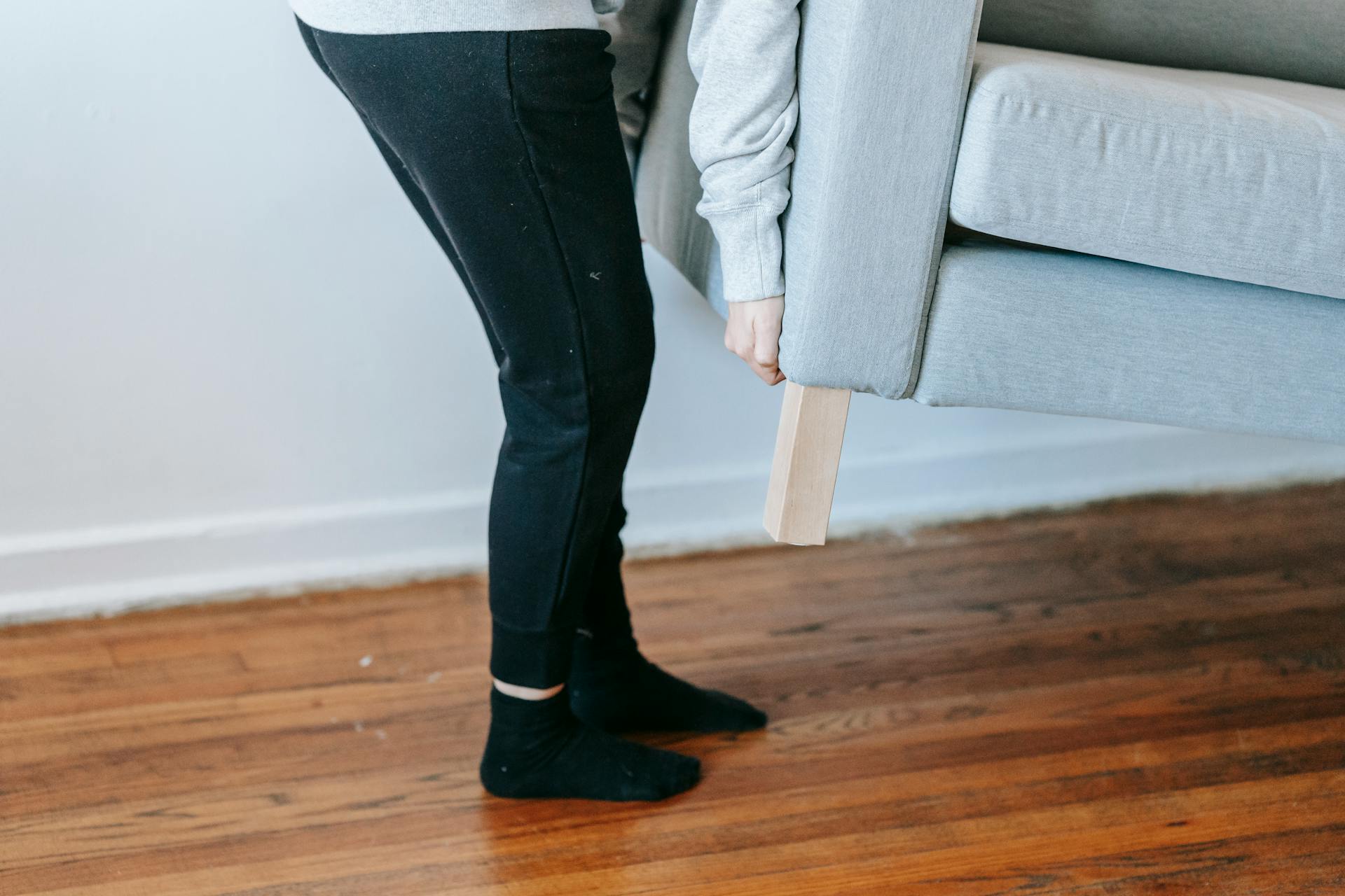 A person lifting a couch in a minimalist living room with wooden floors.