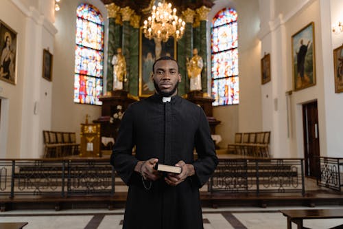 Free Priest in Black Cassock Standing in Front of the Altar Holding a Bible Stock Photo