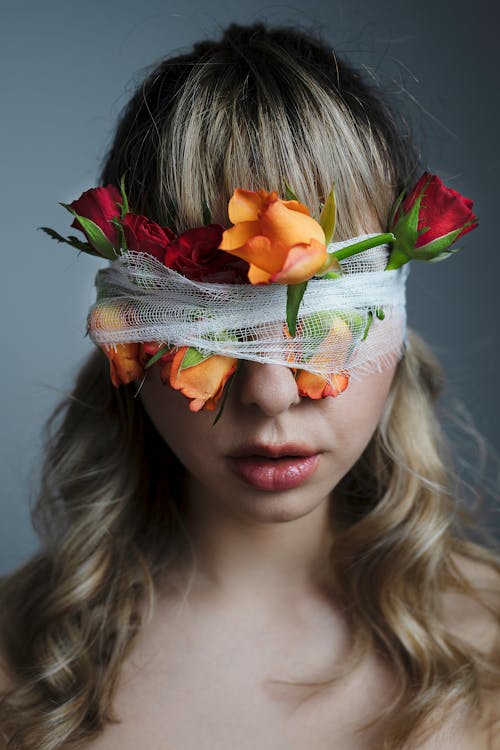 Anonymous female with wavy dyed hair and blindfold with colorful blossoming flowers on gray background