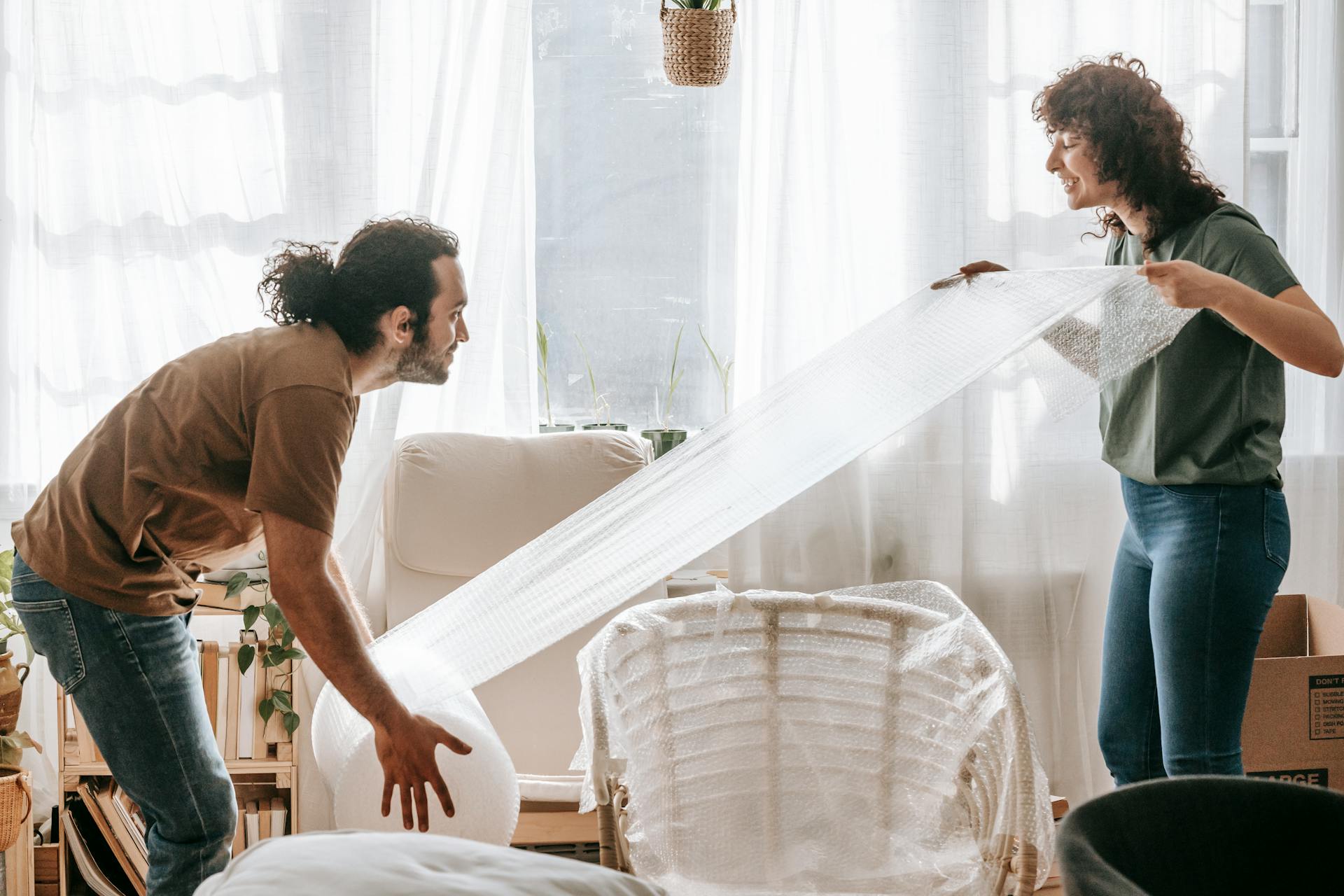 A couple joyfully packs furniture with bubble wrap in their bright living room.
