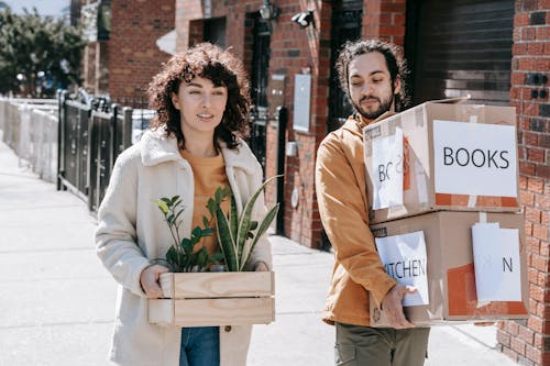 Couple Walking In The Street Carrying Plants And Boxes