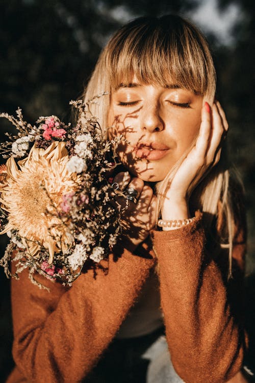 Young mindful female with closed eyes and blossoming flowers touching cheek in soft sunlight on blurred background