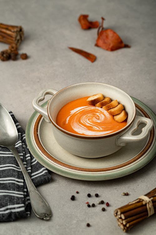 Free Tasty squash puree soup with fresh cream and toasted bread pieces in bowl on table with black peppercorns on gray background Stock Photo