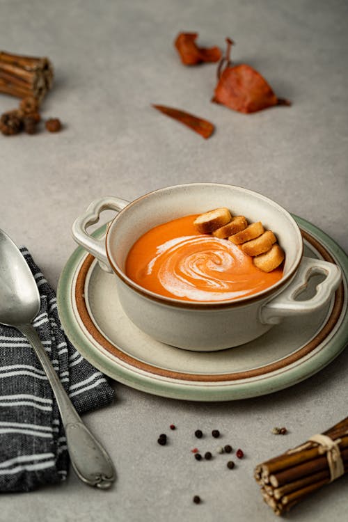 Tasty squash puree soup with fresh cream and toasted bread pieces in bowl on table with black peppercorns on gray background