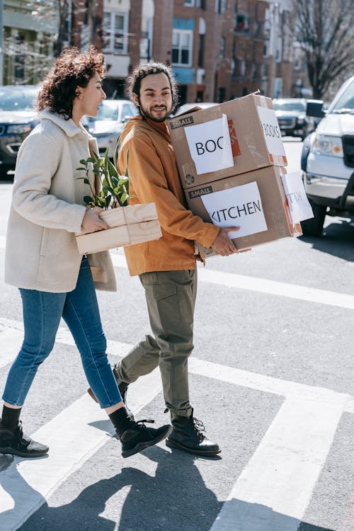 Couple Walking Along The Street Carrying Boxes And Plants