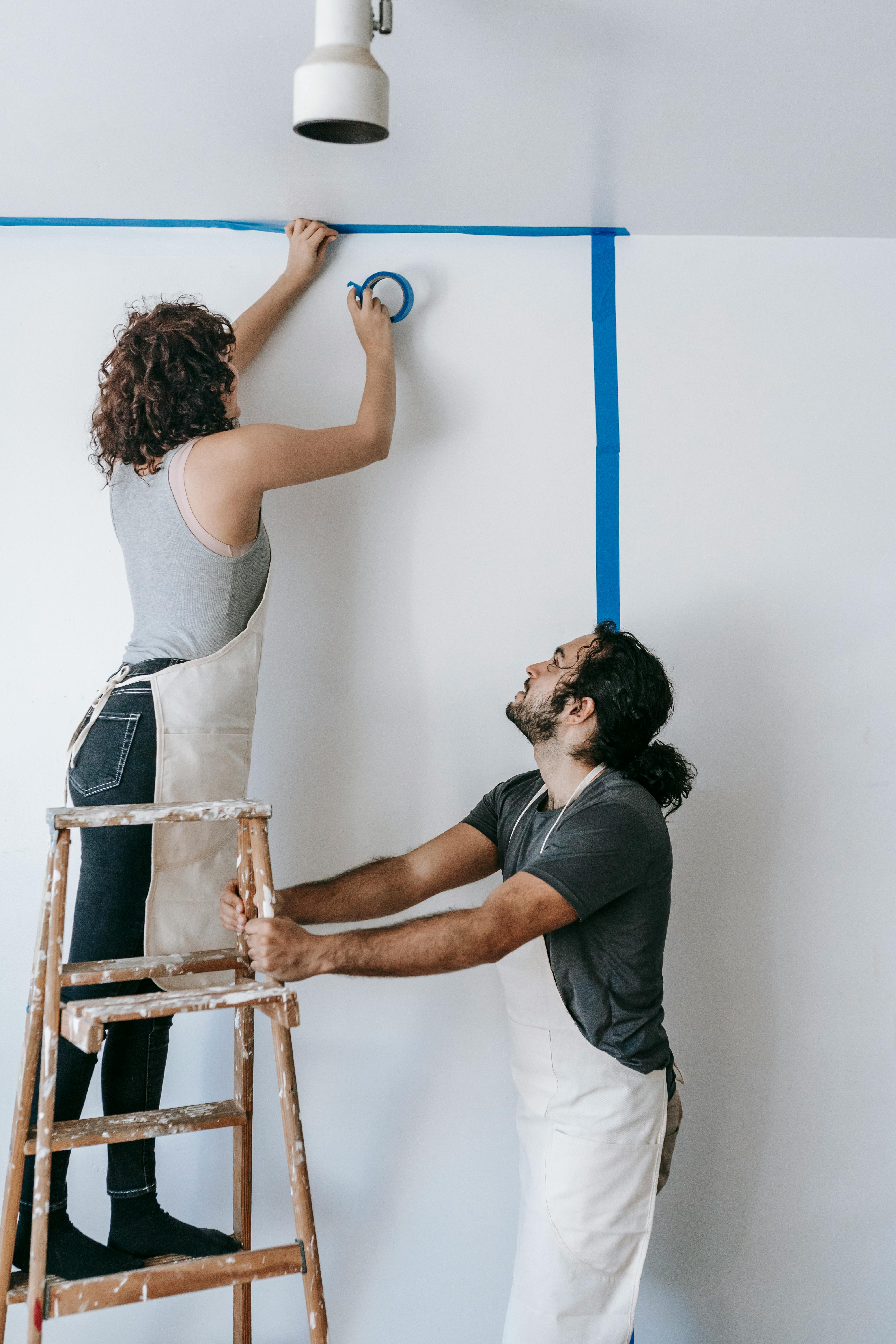 A couple engaged in a home renovation project, using a ladder and tape to prepare walls.