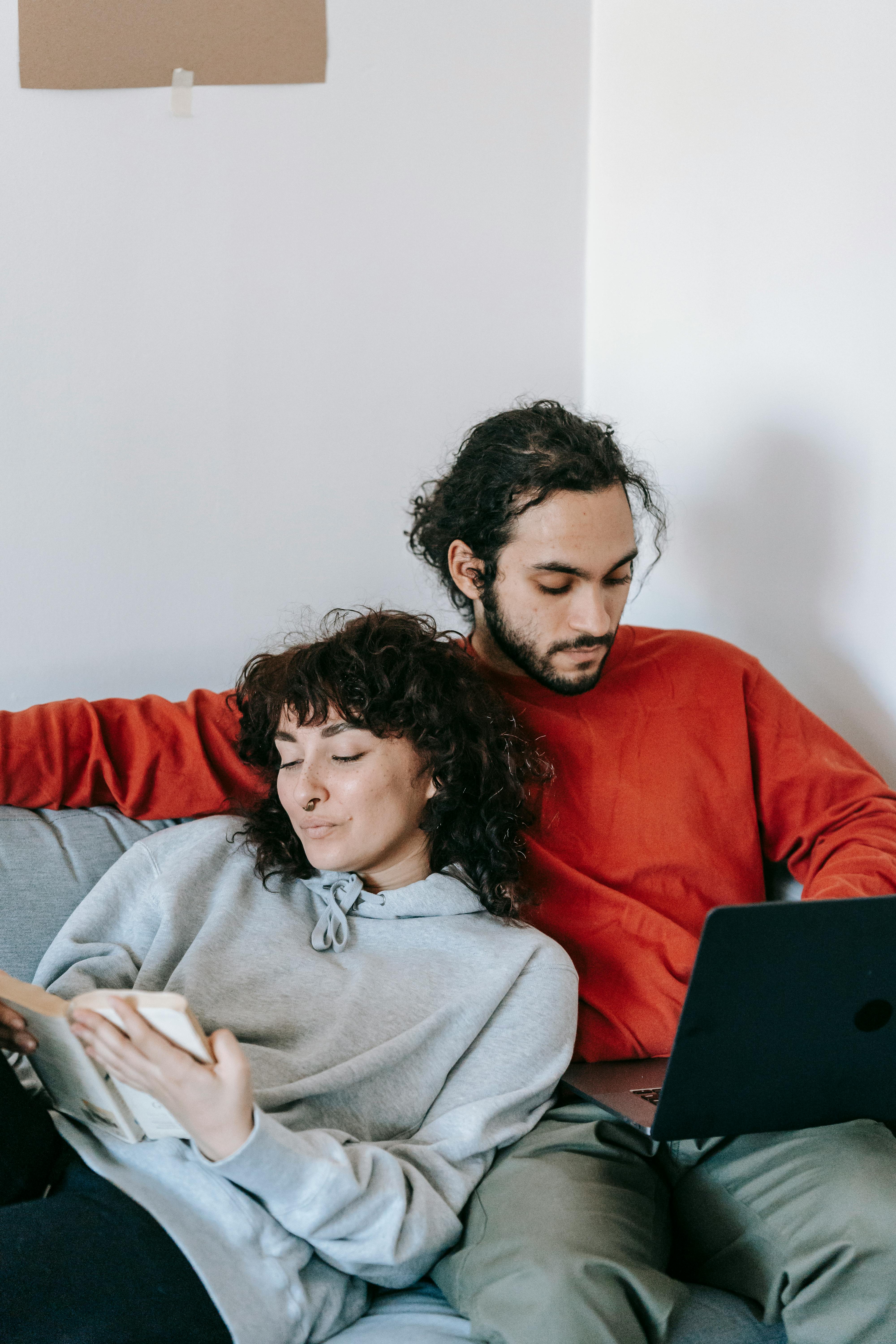 ethnic couple with laptop and book resting on sofa