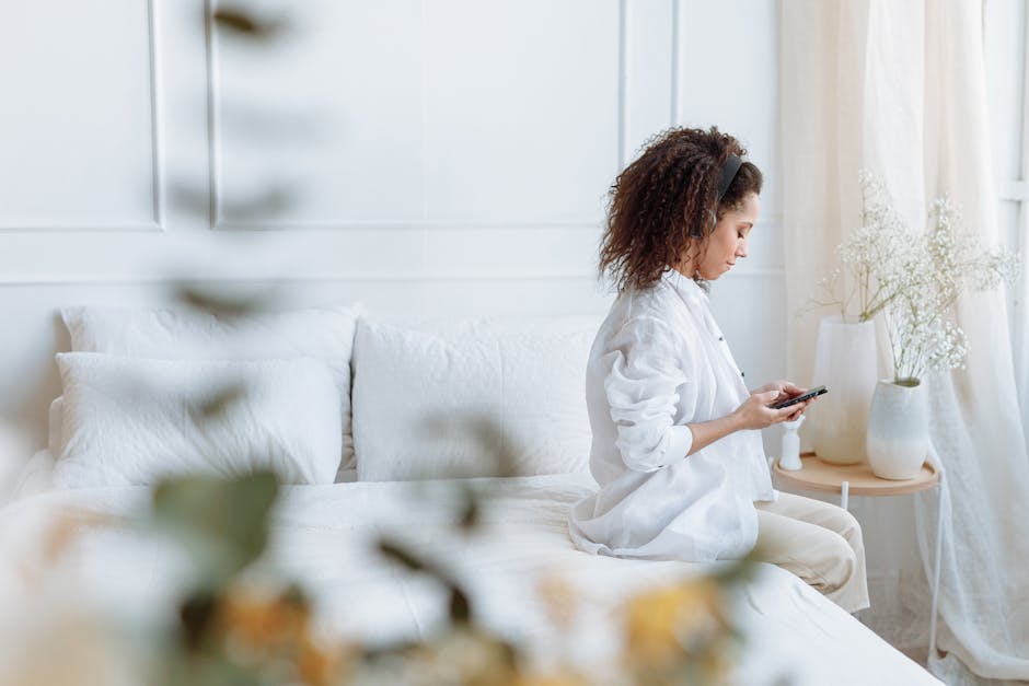 What Sleep Studies Reveal About Adjustable Bed Bases A woman with curly hair sits comfortably on a bed using a smartphone in a bright, cozy bedroom.