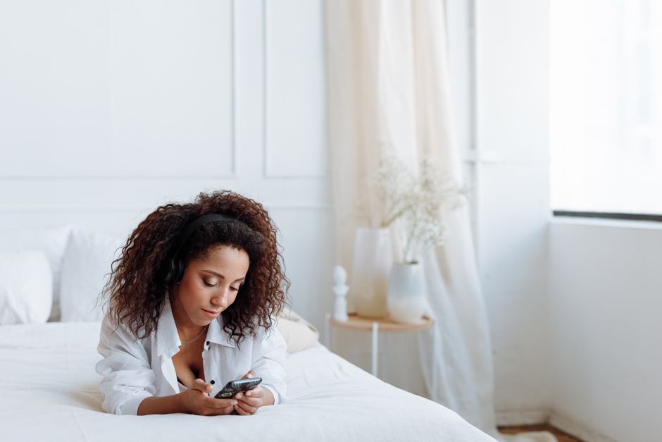 Curly-haired woman in a cozy bedroom using a smartphone with headphones on a white bed.