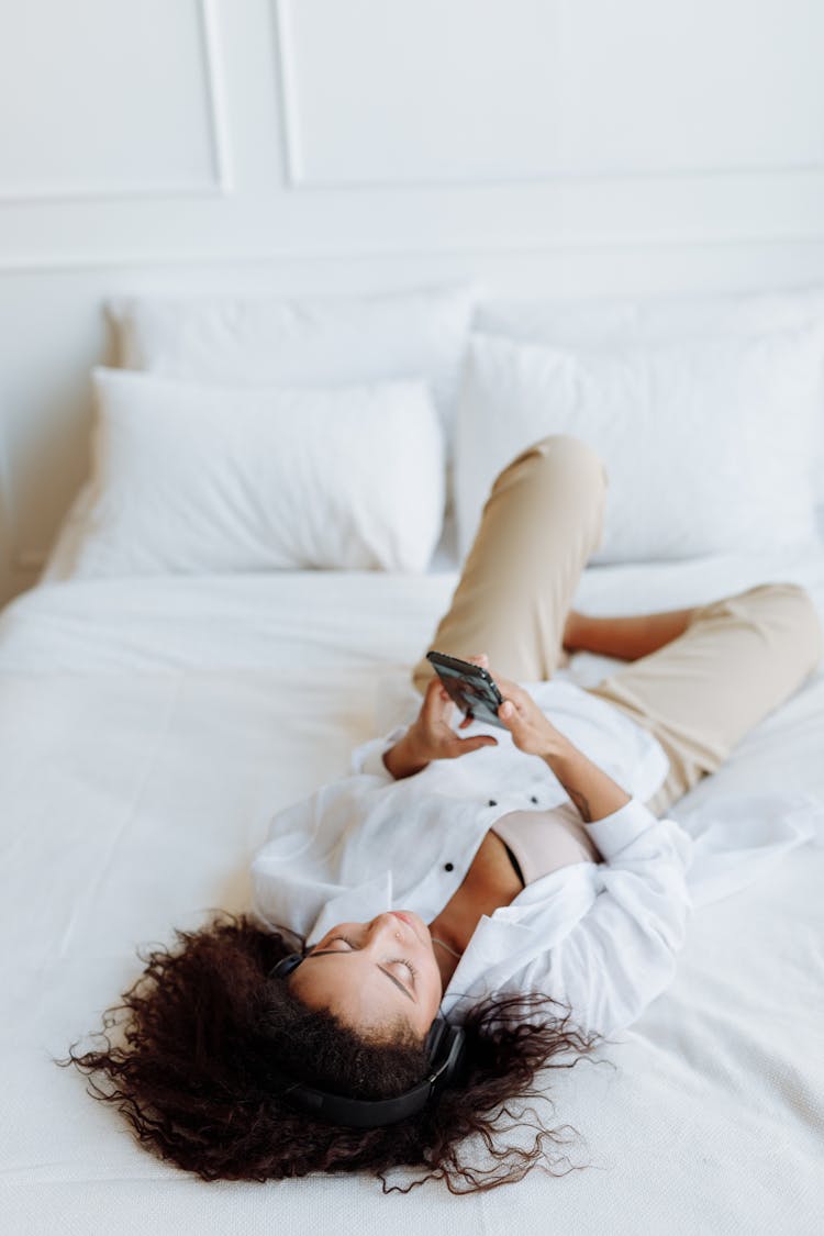 Woman In White Top Lying On Bed While Holding Mobile Phone