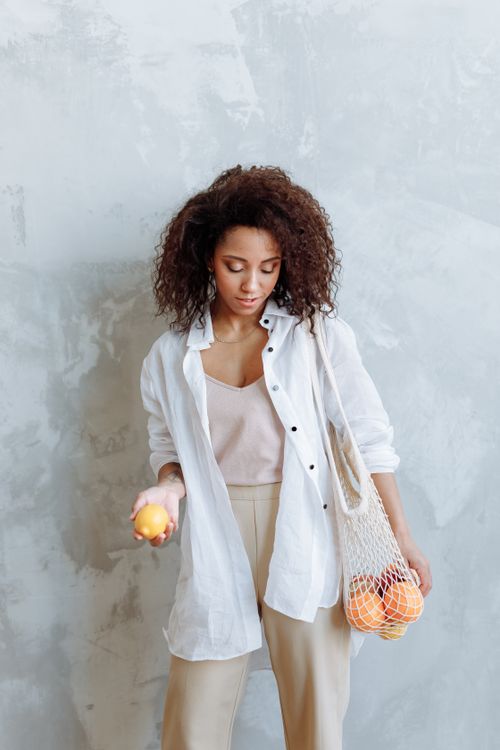 Free Stylish woman with curly hair holding fresh lemons in a net bag against a neutral background. Stock Photo