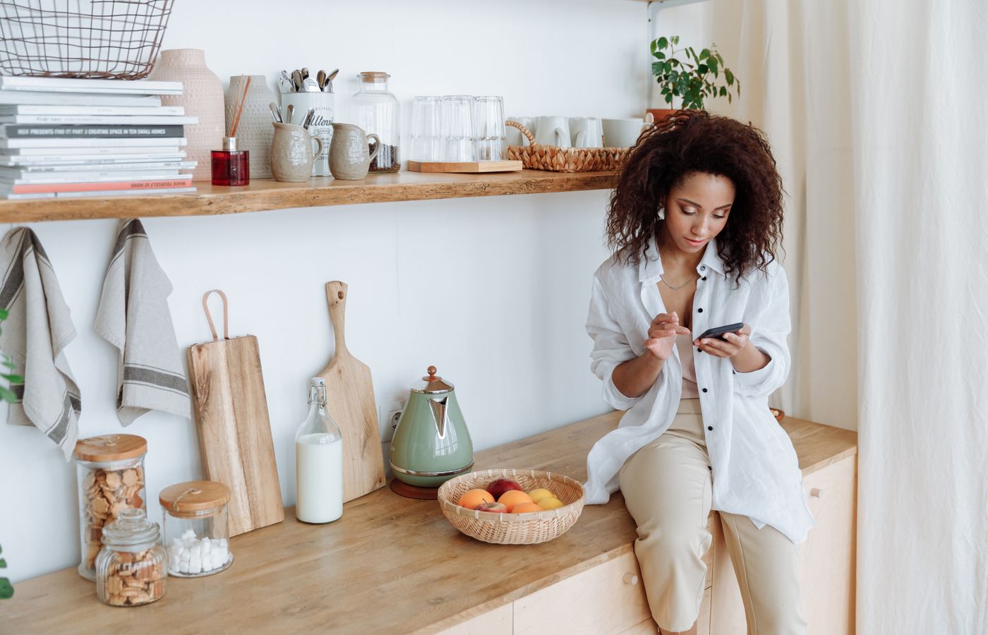 Woman in white shirt browsing smartphone in a cozy kitchen setting with fruits and decor.
