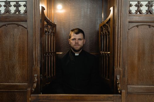 A priest sits solemnly inside a traditional wooden confessional booth.