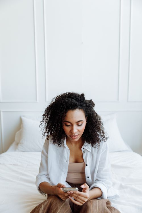 Free A young woman with curly hair using her smartphone while sitting on a bed in a well-lit white room. Stock Photo