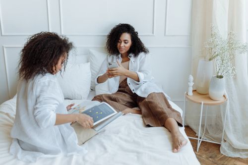 Free Two women sitting on a bed, sharing a relaxing moment with a book and smartphone. Stock Photo