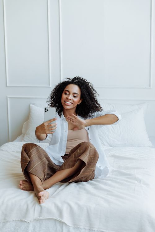 Free A smiling woman with curly hair in a white shirt and brown pants sitting on a bed, taking a selfie. Stock Photo